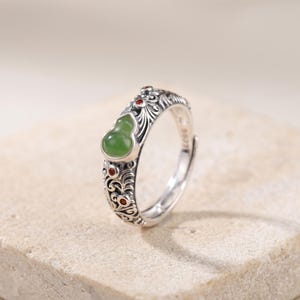 Può includere: Anello d'argento con fascia regolabile, con una pietra verde a forma di zucca di giada e piccoli accenti rossi. L'anello presenta un design ornato e inciso attorno alla fascia.