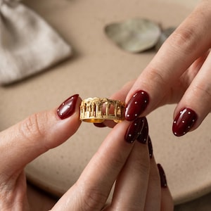 Puede incluir: Un anillo de metal en tono dorado que presenta un dise&ntilde;o de relieve arquitect&oacute;nico en miniatura detallado que envuelve la banda, representando una serie de columnas cl&aacute;sicas y estructuras arqueadas que recuerdan ruinas antiguas.