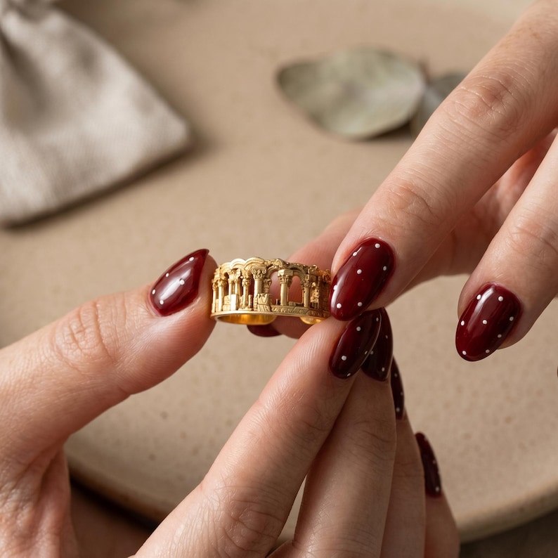 Puede incluir: Un anillo de metal en tono dorado que presenta un dise&ntilde;o de relieve arquitect&oacute;nico en miniatura detallado que envuelve la banda, representando una serie de columnas cl&aacute;sicas y estructuras arqueadas que recuerdan ruinas antiguas.