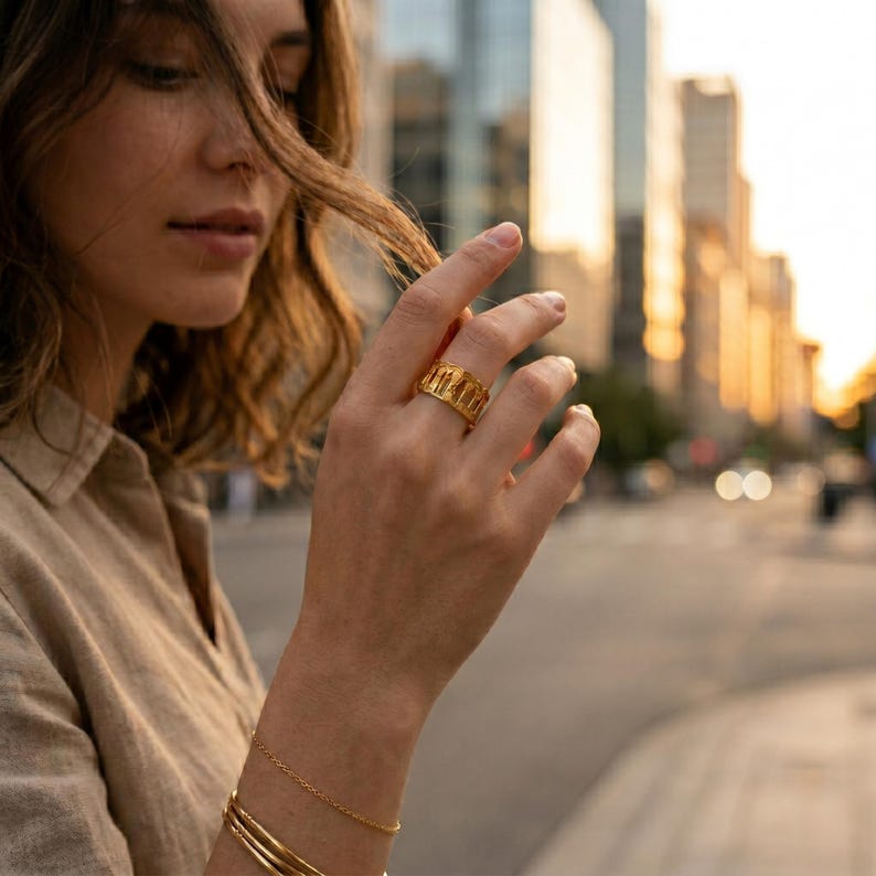 Puede incluir: Un anillo de oro con una serie de cortes verticales alrededor de la banda, usado en el dedo anular. El anillo tiene un acabado pulido y un ancho uniforme. Una pulsera de cadena fina de oro y un brazalete liso de oro se llevan en la misma mu&ntilde;eca.