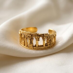 Puede incluir: Un anillo abierto de tono dorado que presenta un dise&ntilde;o en relieve detallado de ruinas arquitect&oacute;nicas antiguas, incluyendo m&uacute;ltiples columnas con capiteles ornamentados y estructuras arqueadas, creando una banda texturizada tridimensional.