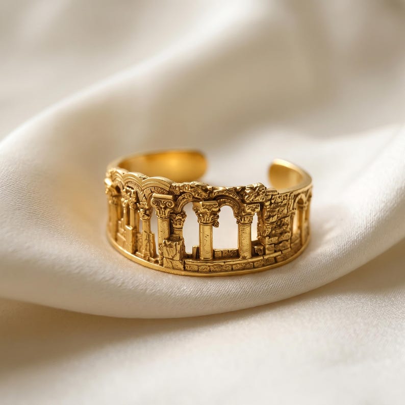 Puede incluir: Un anillo abierto de tono dorado que presenta un dise&ntilde;o en relieve detallado de ruinas arquitect&oacute;nicas antiguas, incluyendo m&uacute;ltiples columnas con capiteles ornamentados y estructuras arqueadas, creando una banda texturizada tridimensional.
