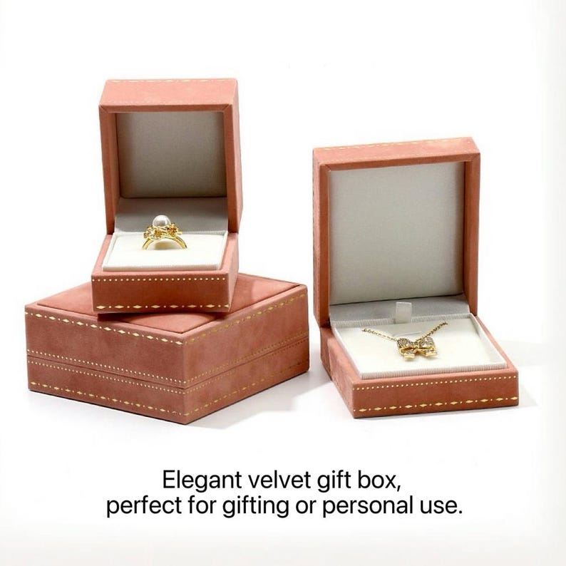 Puede incluir: Dos cajas de joyer&iacute;a de terciopelo rosa empolvado con bordes cosidos en hilo dorado. Una caja cuadrada contiene un anillo dorado con una perla blanca, mientras que la caja rectangular contiene un collar dorado con un dije en forma de lazo.