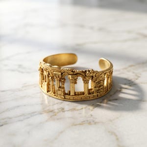 Puede incluir: Un brazalete tipo brazalete de tono dorado con un dise&ntilde;o en relieve detallado de columnas arquitect&oacute;nicas cl&aacute;sicas y ruinas arqueadas. La banda abierta tiene un acabado texturizado y desgastado que imita estructuras de piedra antigua en oro.