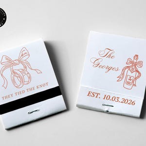 May include: Two white matchboxes with rose-gold illustrations and text. One reads "THEY TIED THE KNOT" with rings and a bow. The other says "The Georges" with a champagne bottle and bow, and "EST. 10.03.2026".