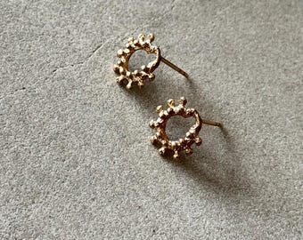 May include: A pair of gold-colored stud earrings. Each earring features a circular design with a textured, beaded appearance. The earrings are displayed on a gray, textured surface.