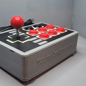 Pode incluir: Um controlador cinza estilo arcade com um joystick vermelho e botões vermelhos e pretos. A superfície superior é preta com detalhes brancos. O texto "TCarterDesigns.com" está impresso na lateral.