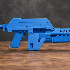 May include: A bright blue toy rifle featuring a blocky, angular design with a prominent number 95 embossed on the side. The toy includes a rear stock, a trigger, a textured grip, and a small orange tip at the end of the barrel with a thin wire.