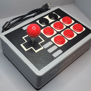 Può includere: Un controller di gioco arcade in stile retrò con un joystick rosso e pulsanti rossi e neri. Il controller ha una combinazione di colori grigio e nero e un cavo USB. Il design ricorda le classiche console di videogiochi.