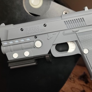 Könnte beinhalten: Eine graue Spielzeugpistole aus Kunststoff mit einem schwarzen Lauf und einem strukturierten Griff. Die Pistole hat einen weißen Knopf an der Seite und die Aufschrift "RETR0 SHOOTER" auf der Oberseite.