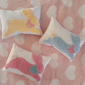 Peut inclure: Trois coussins décoratifs avec des motifs de lapin brodés. Les coussins sont sur une couverture rose et blanche à motifs de cœurs. Les lapins sont jaunes, bleus et roses.