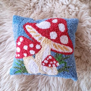May include: A blue throw pillow with a mushroom design. The mushroom is red with white dots and has a white stem with a yellow cap. The pillow is on a white fluffy surface.