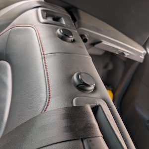 Puede incluir: Primer plano del interior de un asiento de coche. El asiento es de cuero negro con costuras rojas. Se ven un cinturón de seguridad negro y mandos de ajuste. El asiento forma parte del interior de un vehículo.
