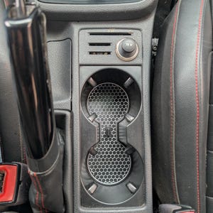 May include: Black car console with a cup holder featuring a honeycomb pattern insert. The console also includes a 12V power outlet and a parking brake lever. The seats are black with red stitching.