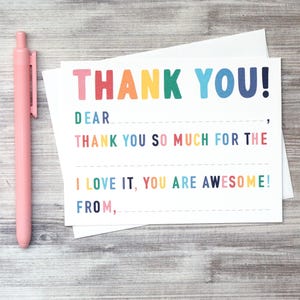 May include: A white thank you card with the words "THANK YOU!" in rainbow colors. The card also includes the phrases "DEAR", "THANK YOU SO MUCH FOR THE", "I LOVE IT, YOU ARE AWESOME!", and "FROM". A pink pen is next to the card.
