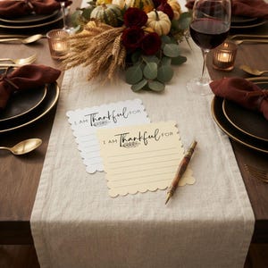 Puede incluir: Dos tarjetas de papel cuadradas con bordes festoneados, una blanca y otra crema, impresas con el texto I AM THANKFUL FOR y l&iacute;neas horizontales para escribir. Un bol&iacute;grafo de madera descansa sobre la tarjeta crema en un camino de mesa beige.