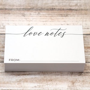 May include: A stack of white notecards with the words "love notes" in elegant script. Below the text is a line, and the word "FROM:" is printed at the bottom. The cards are on a light wood surface.