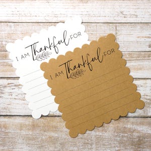 May include: Two scalloped-edge cards, one white and one brown, with the text "I AM Thankful FOR" in black script. The cards have lined spaces for writing and a small leaf design. The cards are on a light wood surface.
