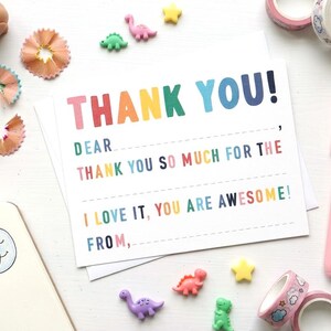 May include: A thank you card with the words "THANK YOU!" in rainbow colours. The card has lines for writing and is surrounded by dinosaur figurines, star-shaped embellishments, and pink washi tape rolls.