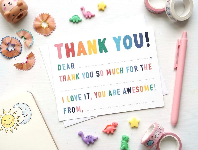 May include: A thank you card with the words "THANK YOU!" in rainbow colours. The card has lines for writing and is surrounded by dinosaur figurines, star-shaped embellishments, and pink washi tape rolls.