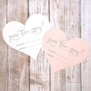 May include: Two heart-shaped cards, one white and one pink, with the text "guess how many?" in elegant script. Each card has lines for "GUESS:" and "NAME:" below the main text, perfect for a party game.