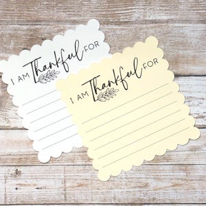 Puede incluir: Dos tarjetas con bordes festoneados, una blanca y otra amarillo p&aacute;lido, cada una con el texto impreso en negro I AM Thankful FOR sobre una peque&ntilde;a ilustraci&oacute;n de una hoja y cinco l&iacute;neas horizontales para escribir notas de gratitud.