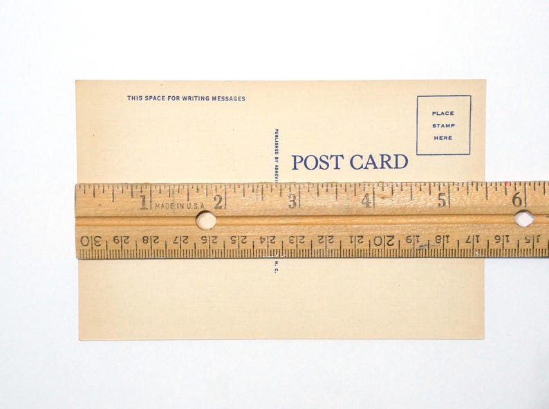 K&ouml;nnte beinhalten: Eine rechteckige beige Postkarte mit blauem Text, der POST CARD, THIS SPACE FOR WRITING MESSAGES und PLACE STAMP HERE lautet. Ein Holzlineal liegt in der Mitte und zeigt, dass die Breite der Postkarte etwa 14 cm betr&auml;gt.