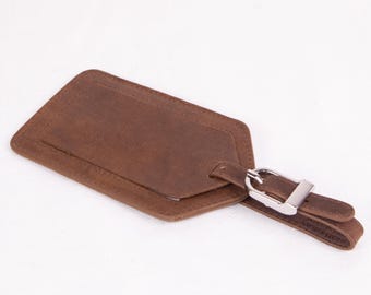 Op de afbeelding: Een bruine leren bagagelabel met een rechthoekige vorm en afgeronde hoeken. De tag heeft een kleiner, verhoogd gedeelte voor een adreskaart en een riem met een zilverkleurige gesp voor bevestiging. Op een witte achtergrond.