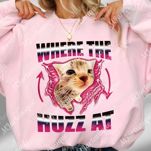 Op de afbeelding: Lichtroze sweatshirt met de tekst "WHERE THE HUZZ AT". De afbeelding toont een katje in een roze bliksemontwerp. De sweatshirt heeft een lichtroze kleur.