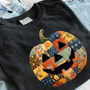 May include: A black t-shirt featuring a large pumpkin graphic composed of various colorful floral and textured fabric patches. The pumpkin design includes black triangular eyes, a triangular nose, and a wide, jagged, black smiling mouth.