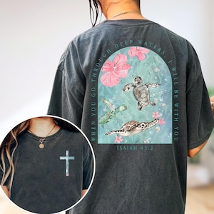Puede incluir: Camiseta gris oscuro con una cruz en la parte delantera y un diseño en la espalda con tres tortugas marinas, flores rosas y el texto "WHEN YOU GO THROUGH DEEP WATERS, I WILL BE WITH YOU" y "ISAIAH 43:2".