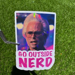 Pode incluir: Um autocolante com o retrato de um homem com óculos e o texto rosa "GO OUTSIDE NERD". O autocolante tem um efeito holográfico e está sobre um fundo verde.