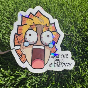 Pode incluir: Um autocolante holográfico com uma personagem de desenho animado com olhos arregalados e boca aberta. A personagem tem cabelo amarelo e detalhes laranja. O texto no autocolante diz: "WHAT THE HELL IS THAT?!?!"