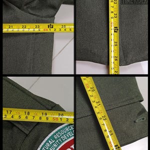 Pode incluir: Fotos em close-up de uma jaqueta verde-escura com um patch que diz "NATURAL RESOURCES COMMUNITY DEVELOPMENT". A jaqueta &eacute; medida com uma fita m&eacute;trica amarela, mostrando um comprimento de aproximadamente 63,5 cm. A marca "TIME RIDER" &eacute; vis&iacute;vel.