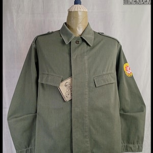 Puede incluir: Chaqueta verde oliva con dos bolsillos en el pecho y cuello, exhibida en un maniquí. Un parche con un emblema rojo y amarillo está en la manga izquierda. Una etiqueta con un logotipo está adherida a la chaqueta.