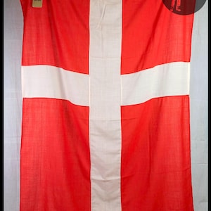 Könnte beinhalten: Eine große rote Flagge mit einem weißen Kreuz. Die Flagge ist aus gewebtem Stoff und hat eine leicht strukturierte Oberfläche.