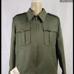 Pode incluir: Casaco verde-oliva com fecho de correr, dois bolsos no peito e dois bolsos laterais. O casaco tem gola e ombreiras. Uma etiqueta com um log&oacute;tipo est&aacute; anexada. O casaco &eacute; exibido num manequim.