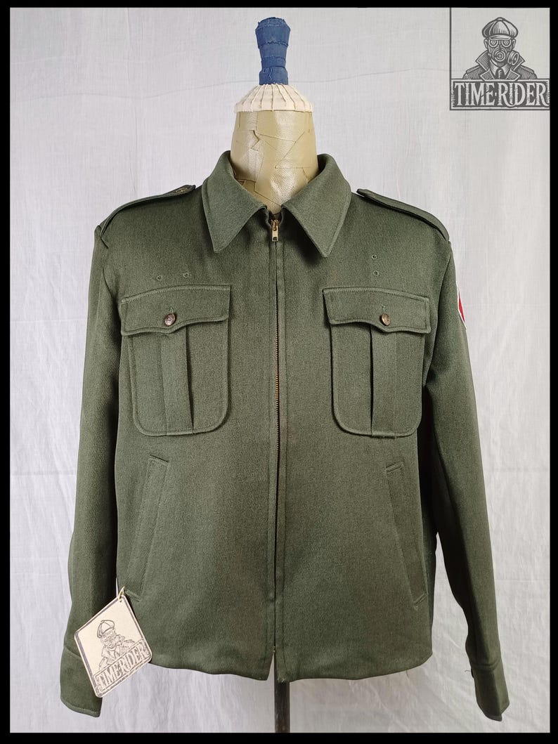 Pode incluir: Casaco verde-oliva com fecho de correr, dois bolsos no peito e dois bolsos laterais. O casaco tem gola e ombreiras. Uma etiqueta com um log&oacute;tipo est&aacute; anexada. O casaco &eacute; exibido num manequim.