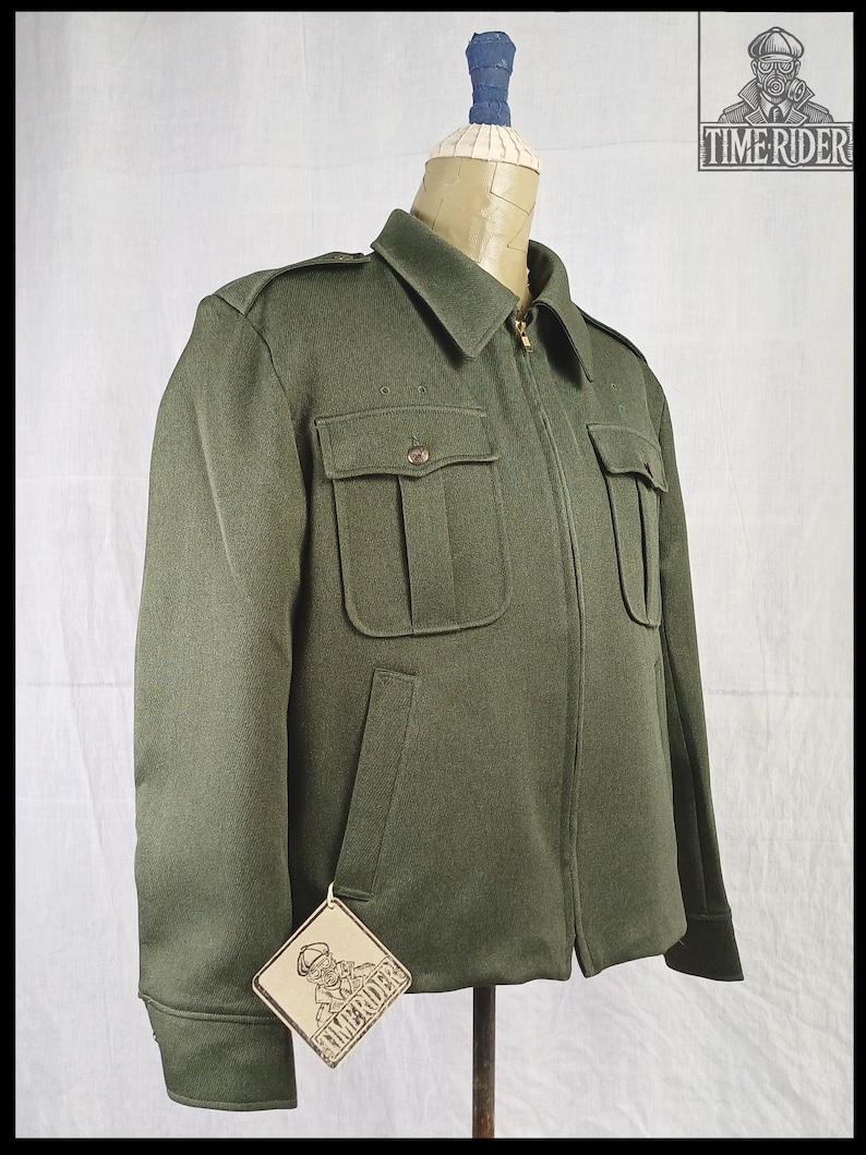Pode incluir: Casaco verde-oliva com fecho de correr, dois bolsos no peito e dois bolsos laterais. O casaco tem gola e ombreiras. Uma etiqueta com o texto "TIMERIDER" est&aacute; presa ao casaco.