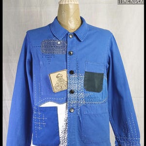 Puede incluir: Chaqueta azul con parches y costuras visibles. La chaqueta tiene cuello, cierre de botones y múltiples bolsillos. Una etiqueta de madera con el texto "TIME RIDER" está adjunta. La chaqueta se exhibe en un maniquí.