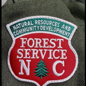 Pode incluir: Um patch do Servi&ccedil;o Florestal com formato de escudo vermelho, borda branca e texto verde e branco. A faixa superior diz "NATURAL RESOURCES AND COMMUNITY DEVELOPMENT". O centro diz "FOREST SERVICE N C" com um gr&aacute;fico de &aacute;rvore verde.