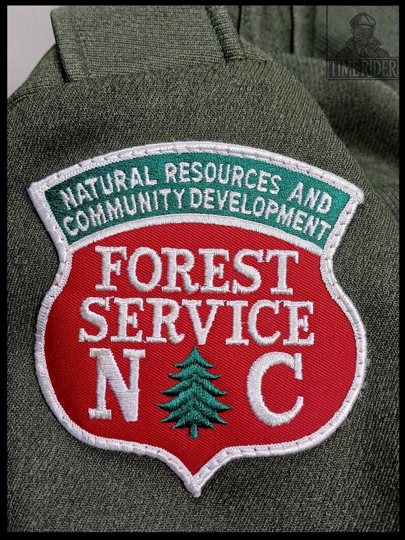 Pode incluir: Um patch do Servi&ccedil;o Florestal com formato de escudo vermelho, borda branca e texto verde e branco. A faixa superior diz "NATURAL RESOURCES AND COMMUNITY DEVELOPMENT". O centro diz "FOREST SERVICE N C" com um gr&aacute;fico de &aacute;rvore verde.