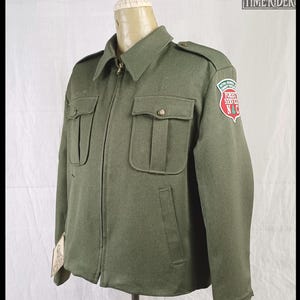 Pode incluir: Casaco verde-oliva com fecho de correr, dois bolsos no peito e dois bolsos inferiores. O casaco tem gola e ombreiras. Um patch na manga diz "Forest Service N&C". O casaco &eacute; exibido em um manequim.