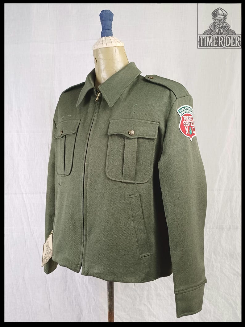 Pode incluir: Casaco verde-oliva com fecho de correr, dois bolsos no peito e dois bolsos inferiores. O casaco tem gola e ombreiras. Um patch na manga diz "Forest Service N&C". O casaco &eacute; exibido em um manequim.