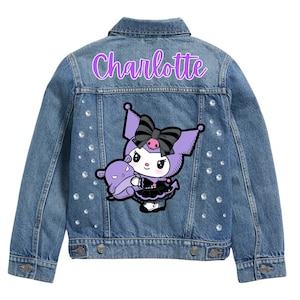 Op de afbeelding: Een lichtblauwe spijkerjas met de naam "Charlotte" in paars script. De achterkant heeft een stripfiguur met een zwarte strik en een paars wezen. Strass-steentjes zijn verspreid over de achterkant.