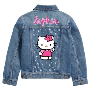 Op de afbeelding: Blauwe spijkerjas met een Hello Kitty-ontwerp en de naam "Sophia" in roze letters. De jas heeft strass-details en klassieke spijkerjasdetails zoals knoopsluitingen en zakken.