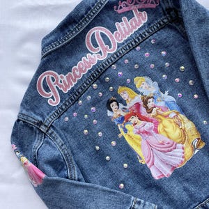 Op de afbeelding: Een blauwe denimjas met een roze glitterpatch waarop "Princess Delilah" staat en een kleurrijk beeld van Disney-prinsessen op de achterkant. De jas is versierd met iriserende strasssteentjes.