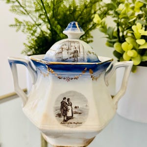 May include: A white and blue porcelain sugar bowl with a lid. The lid and rim feature a blue band with gold accents. The bowl has two handles and a decorative image of a couple. The text "Return of the Mayflower" is below the image.