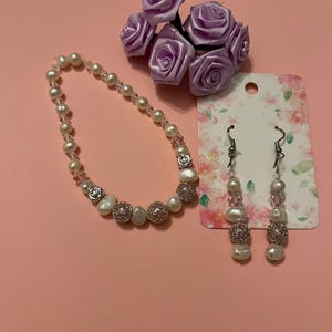 May include: A pearl necklace and matching earrings set with silver accents and a cluster of purple fabric roses. The jewelry is displayed on a pink surface, showcasing the delicate design and craftsmanship.