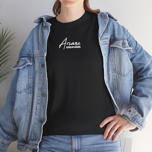 May include: Black t-shirt with the white text "Arcane LEAGUE OF LEGENDS" printed on the front. The shirt is worn with a blue denim jacket and blue jeans.
